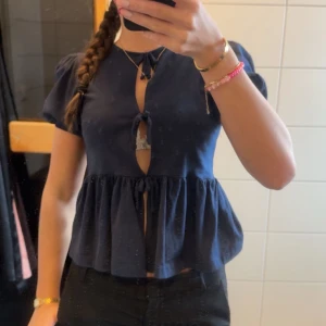 Mörkblå blus med knytning - Säljer en söt mörkblå blus med knytning framtill och volangdetaljer. Blusen har korta ärmar och en luftig passform, perfekt för varmare dagar. 