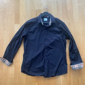Svart skjorta från Burberry - Snygg svart skjorta från Burberry med klassiskt rutigt mönster på insidan av kragen och ärmsluten. Skjortan har långa ärmar och knappar framtill. Perfekt för en stilren look. Är storlek L men passar som ”M” mot den mindre sidan.