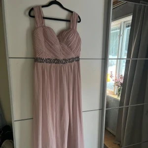 Ljusrosa långklänning från Little Mistress - Elegant ljusrosa långklänning från Little Mistress med breda axelband och vackert draperad överdel. Klänningen har en glittrig midjedetalj som ger en extra touch av glamour. Perfekt för speciella tillfällen.
