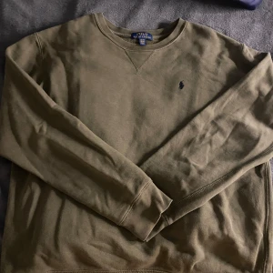 Olivgrön tröja från Polo Ralph Lauren - Säljer en olivgrön tröja från Polo Ralph Lauren med rund halsringning och broderad logotyp på bröstet. Tröjan har långa ärmar och är perfekt för en avslappnad stil. Passar bra till både jeans och chinos.