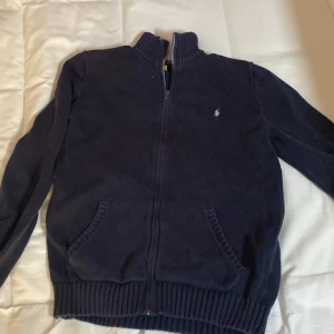Mörkblå stickad tröja med dragkedja från Polo Ralph Lauren - Säljer en mörkblå stickad tröja från Polo Ralph Lauren med dragkedja och broderad logga på bröstet. Tröjan har långa ärmar och ribbade muddar vid ärmslut och nederkant. Perfekt för kyliga dagar! Ordinarie storlek: XL Storlek nu:S