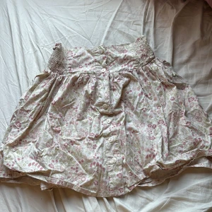 Blommig kjol med knappar - Charmig kortkjol med blommigt mönster i rosa och gröna nyanser. Kjolen har en elastisk midja och knappar framtill för en söt och bekväm stil. Det saknas en knapp men inget man märker av tydligt.