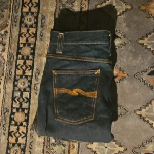 Mörk blåa jeans från Nudie Jeans - Snygga svarta jeans från Nudie Jeans med orange sömmar och klassisk femficksdesign. Jeansen har en rak passform och är tillverkade i denim. Perfekta för en stilren look. Messa vid minsta fundering🙏