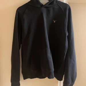 Svart hoodie från GANT - Säljer en stilren svart hoodie från GANT med deras klassiska logga på bröstet. Tröjan har en bekväm passform med långa ärmar och en praktisk huva. Perfekt för en avslappnad look. Storlek 176cm / Xs
