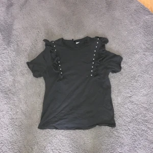 Svart topp med volangdetaljer - Snygg svart topp med volangdetaljer och pärlor på axlarna. Perfekt för en stilren look med det lilla extra. Toppen har korta ärmar och en normal passform.