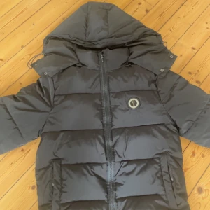 Svart pufferjacka från Trapstar - Säljer en svart pufferjacka från Trapstar med huva och dragkedja. Jackan har ett broderat Trapstar-märke på ryggen och en rund logotyp på bröstet. Perfekt för kyliga dagar. Använd 1 gång