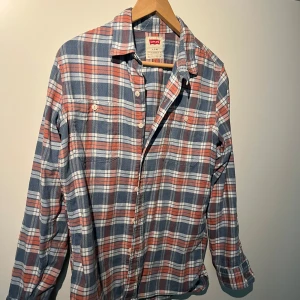 Rutig skjorta från Levi's - Säljer en snygg rutig skjorta från Levi's i blått, vitt och rött. Skjortan har långa ärmar och knappar framtill. Perfekt för en avslappnad stil. Tillverkad i ett mjukt och bekvämt material.