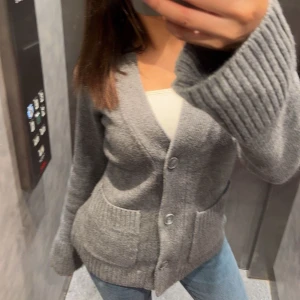 Grå kofta med knappar från Åhléns  - Säljer en mysig grå kofta med knappar framtill och två fickor. Koftan har långa ärmar och ribbade muddar, perfekt för kyliga dagar. Passar bra till jeans eller kjol.