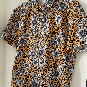 Mönstrad kortärmad skjorta - Säljer en cool kortärmad skjorta med ett unikt mönster av blommor och leopardfläckar i svart och vitt på en gul bakgrund. Perfekt för att sätta statement, testa med ett par svarta kostymbyxor eller svarta jeans för en elevated look! Nypris: 699kr. 