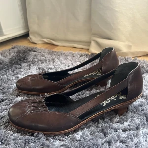 Bruna ballerinaskor från Rieker - Snygga bruna ballerinaskor från Rieker med låg klack och dekorativ rynkning framtill. Skorna har en öppen design på sidorna och är tillverkade i skinn. Perfekta för en stilren look.
