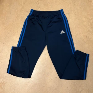 Svarta träningsbyxor från Adidas - Säljer ett par svarta träningsbyxor från Adidas med blå ränder längs sidorna. Byxorna har en elastisk midja och är perfekta för sport och träning. Adidas-loggan är tryckt på framsidan.