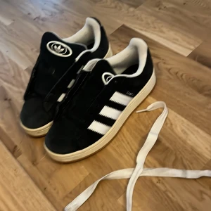 Svarta sneakers från Adidas - Snygga svarta sneakers från Adidas med de klassiska tre vita ränderna på sidan. Skorna har en vit sula och vita snören, vilket ger en stilren kontrast. Perfekta för en avslappnad stil.