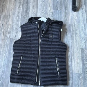 Svart dunväst från Moose Knuckles - Snygg svart dunväst från Moose Knuckles med dragkedja och huva. Västen har en quiltad design och två praktiska fickor med dragkedjor. Perfekt för kyliga dagar när du vill hålla stilen.