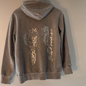 Grå hoodie med paljettvingar från Moocci - Säljer en grå hoodie från Moocci med en cool detalj av paljettvingar på ryggen. Tröjan har en klassisk känguruficka och justerbar huva med dragsko. Perfekt för en avslappnad stil med en touch av glitter. ✨ vingarna liknar Victorias secret 💘