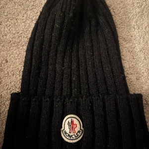 Svart ribbad mössa från Moncler - Snygg svart ribbad mössa från Moncler med broderad logotyp framtill. Perfekt för att hålla värmen under kyliga dagar. Stilren design som passar till det mesta.