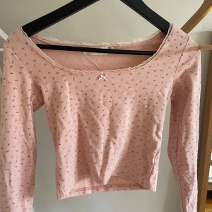 Rosa blommig topp med spetsdetaljer - Söt rosa topp med små blommor och spetsdetaljer vid halsringningen. Toppen har en liten rosett framtill och långa ärmar. Perfekt för en romantisk look.