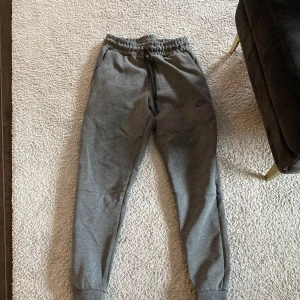 Grå mjukisbyxor från Nike - Säljer ett par grå mjukisbyxor från Nike med elastisk midja och snörning. Byxorna har en avslappnad passform och är perfekta för en chill dag. Diskret Nike-logga på benet.