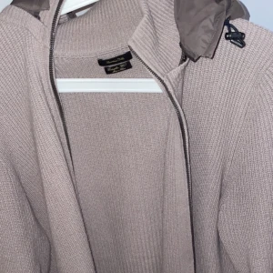  stickad jacka från Massimo Dutti - Säljer en stilren  stickad jacka från Massimo Dutti. Jackan har en dragkedja framtill och en huva i kontrasterande material. Perfekt för kyliga dagar när du vill hålla stilen. Tillverkad i extrafin ull för en lyxig känsla. Huvan går att ta av med hjälp av dragkedja