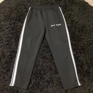  Palm Angels byxor - Snygga svarta byxor från Palm Angels med vita ränder längs sidorna och logotyp på framsidan. Byxorna har en elastisk midja och har en bekväm passform. finns en jätteliten fläck på framsidan men inget som lägger märke till eller syns vid användning. Hör av dig om du har några funderingar.