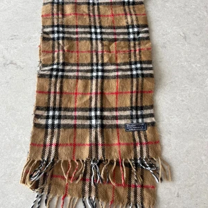Rutig halsduk från Burberry - Säljer en klassisk rutig halsduk från Burberry i 100% lammull. Den har en beige bas med svarta, vita och röda rutor samt fransar i ändarna. NYSKICK, skriv fall du har frågor eller funderingar!❤️🖤