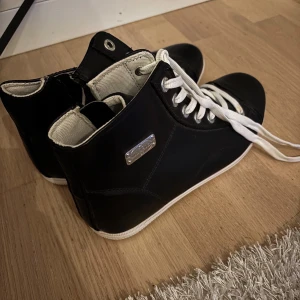 Svarta sneakers med snörning - Snygga svarta sneakers med vita snören och en metallplatta på sidan. Skorna har en högre modell med rund tå och vit sula. Perfekta för en stilren look. Märke: vox shoes. Aldrig använda 