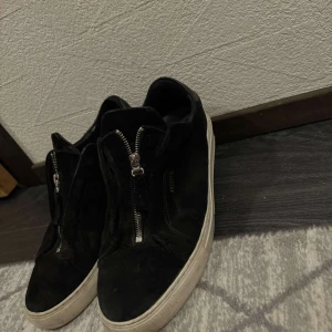 Arigato skor - Snygga svarta arigatos i mocka med dragkedja framtill. Perfekta för en stilren look med en bekväm passform. Den vita sulan ger en fin kontrast till det svarta.