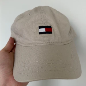 Beige keps från Tommy Hilfiger - Snygg beige keps från Tommy Hilfiger med justerbart spänne baktill. Kepsen har en broderad logotyp framtill i rött, vitt och blått. Perfekt för en avslappnad stil.