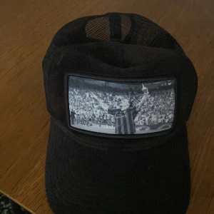 Svart keps från Berretto Sweden - Snygg svart keps från Berretto Sweden med ett tryck av en fotbollsspelare på framsidan. Kepsen har ett nätmaterial baktill för bättre ventilation och justerbar passform. Perfekt för fotbollsfans! Den är i nyskick 