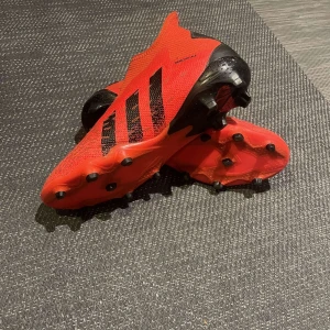Röda fotbollsskor från Adidas - Snygga röda fotbollsskor från Adidas, modellen predator med svarta detaljer och dobbar för optimalt grepp på planen. Skorna har en strömlinjeformad design utan snörning för en tight passform och maximal komfort. Säljs inte längre. Storlek 44. Inget medföljer🙃Hör av er vid frågor😁😁