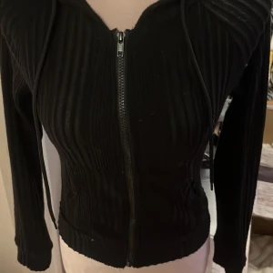 Svart ribbad hoodie med dragkedja - Snygg svart hoodie med ribbad struktur och dragkedja framtill. Den har en luva med dragsko och långa ärmar. Perfekt för en casual look. Storlek M men skulle säga att den sitter som en S