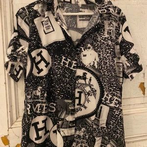 Svartvit mönstrad skjorta/tunika stl S/M  - Säljer en unik svartvit skjorta/tunika med ett iögonfallande mönster av olika logotyper och motiv. Skjortan har korta ärmar och knappar framtill. Perfekt för den som vill sticka ut med en djärv stil.