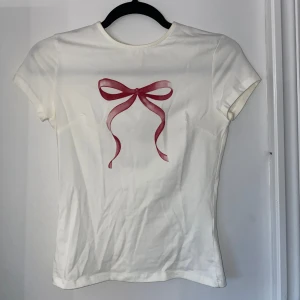 Vit t-shirt med rosett - Säljer en söt vit t-shirt med en röd/rosa rosett på framsidan. T-shirten har korta ärmar och en normal passform. Perfekt för en avslappnad stil! Extremt skönt material och bekväm passform.
