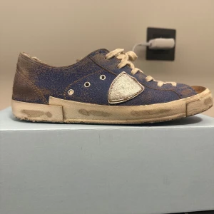 Blå och bruna sneakers från Philippe Model - Snygga sneakers i blått och brunt från Philippe Model. Skorna har en unik design med snörning och en distinkt logga på sidan. Perfekta för en avslappnad stil.