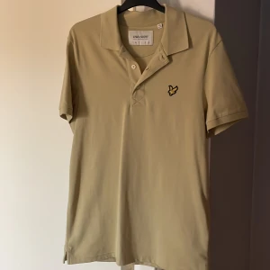 Beige pikétröja från Lyle & Scott - Säljer en stilren beige pikétröja från Lyle & Scott. Tröjan har en klassisk krage med knappar och det ikoniska logotypmärket på bröstet. Perfekt för en avslappnad men ändå snygg look.