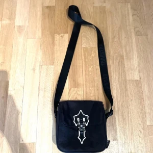 Svart axelväska från Trapstar - Snygg svart axelväska från Trapstar med ett unikt spänne framtill. Väskan har en justerbar axelrem med märkeslogga och en praktisk dragkedja för säker förvaring. Perfekt för att bära med sig det nödvändigaste med stil.