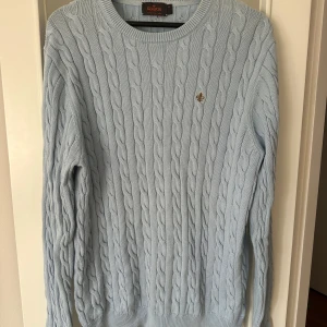 Ljusblå kabelstickad tröja från Morris - Säljer en stilren ljusblå kabelstickad tröja från Morris. Tröjan har en klassisk rund halsringning och ett diskret emblem på bröstet. 