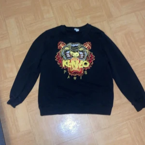 Svart sweatshirt från Kenzo - Säljer en svart sweatshirt från Kenzo med ett stort, färgglatt tigerbroderi på framsidan. Tröjan har långa ärmar och en rund halsringning. Den är XS det är barn storlek. Perfekt för en cool och avslappnad stil. Hör av er för minsta lilla✅