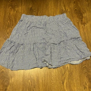 Blå mönstrad kjol - Säljer en söt blå kortkjol med vitt mönster. Kjolen har en volangdetalj och elastisk midja för en bekväm passform. Perfekt för varma dagar!