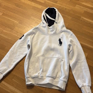 Vit hoodie från Polo Ralph Lauren - Säljer en stilren vit hoodie från Polo Ralph Lauren med den klassiska loggan broderad på bröstet. Hoodien har en känguruficka och justerbar huva med dragsko. Perfekt för en avslappnad stil.