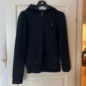 Blå hoodie från Ralph Lauren - Säljer en stilren mörkblå hoodie från Ralph Lauren med dragkedja och det klassiska logotypbroderiet på bröstet. Perfekt för en avslappnad look. Den har långa ärmar och en praktisk huva.