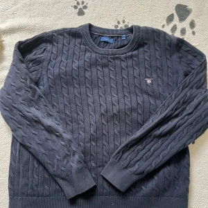 Mörkblå kabelstickad tröja från GANT - Säljer en stilren mörkblå kabelstickad tröja från GANT. Tröjan har en rund halsringning och långa ärmar, perfekt för kyligare dagar. Den klassiska designen gör den lätt att matcha med olika outfits. Passar både till vardag och mer uppklädda tillfällen.