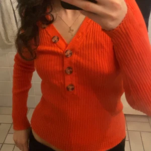 Röd ribbad tröja med knappar - Säljer en snygg orange/ röd ribbad tröja med stora knappar framtill. Tröjan har en v-ringad design och långa ärmar, perfekt för en stilren look. Passar bra till både jeans och kjol. Passar s också