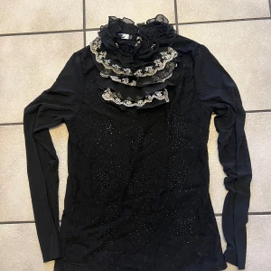 Svart långärmad blus med spetsdetaljer storlek S/M - Elegant svart blus från Bik Bok med långa ärmar och vackra spetsdetaljer. Blusen har en unik design med volanger och broderade spetsar i halsen. Perfekt för en stilfull look.