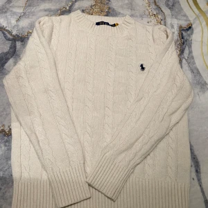 Vit stickad tröja från Polo Ralph Lauren - Säljer en stilren vit stickad tröja från Polo Ralph Lauren med klassiskt flätmönster. Tröjan har långa ärmar och en broderad logotyp på bröstet. Perfekt för kyliga dagar!