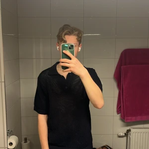Svart pikétröja från Massimo Dutti - Snygg svart pikétröja från Massimo Dutti med korta ärmar och klassisk krage. Tröjan har en unik stickad design som ger en luftig känsla. Perfekt för en stilren look.