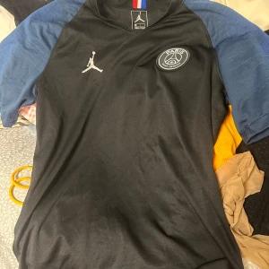 Svart fotbollströja från Jordan x PSG - Snygg svart fotbollströja från Jordan i samarbete med PSG. Tröjan har korta ärmar i blått och ett stilrent PSG-emblem på bröstet. Perfekt för fotbollsälskare som vill ha något unikt och sportigt i garderoben. S-m