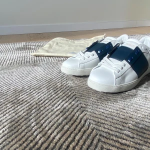 Vita sneakers med blå detalj - Snygga vita sneakers från Valentino med en unik blå rem över snörningen. Skorna har en klassisk rund tå och är dekorerade med de ikoniska nitarna baktill. Perfekta för en stilren look.
