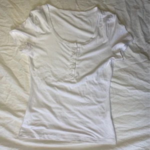 Vit kortärmad topp med knappar - Säljer en vit kortärmad topp med rund halsringning och knappar framtill. Perfekt för en avslappnad stil. Passar bra till jeans eller kjol.