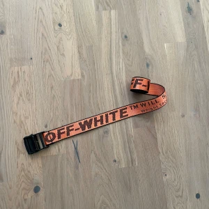 Orange bälte från Off-White - Säljer ett coolt orange bälte från Off-White med svart text och en robust spänne. Perfekt för att ge din outfit en edgy touch. Bältet har en industriell design och är justerbart för bästa passform.