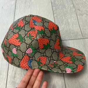 Gucci jordgubbs keps  - Snygg keps från Gucci med ett lekfullt jordgubbsmönster i rött och grönt på en beige bakgrund. Kepsen har en justerbar rem i rosa på baksidan och en rosa insida. Perfekt för att ge din outfit en färgglad touch!